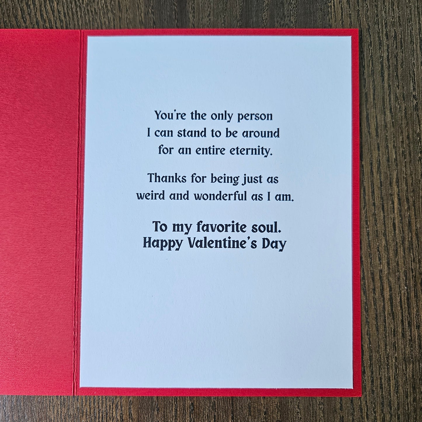 A square red greeting card with a white insert featuring printed black text, photographed from above on a dark wood surface.