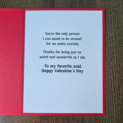 A square red greeting card with a white insert featuring printed black text, photographed from above on a dark wood surface.