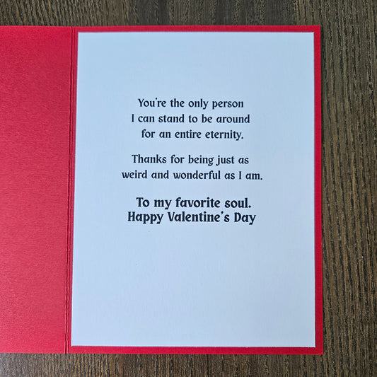 A square red greeting card with a white insert featuring printed black text, photographed from above on a dark wood surface.