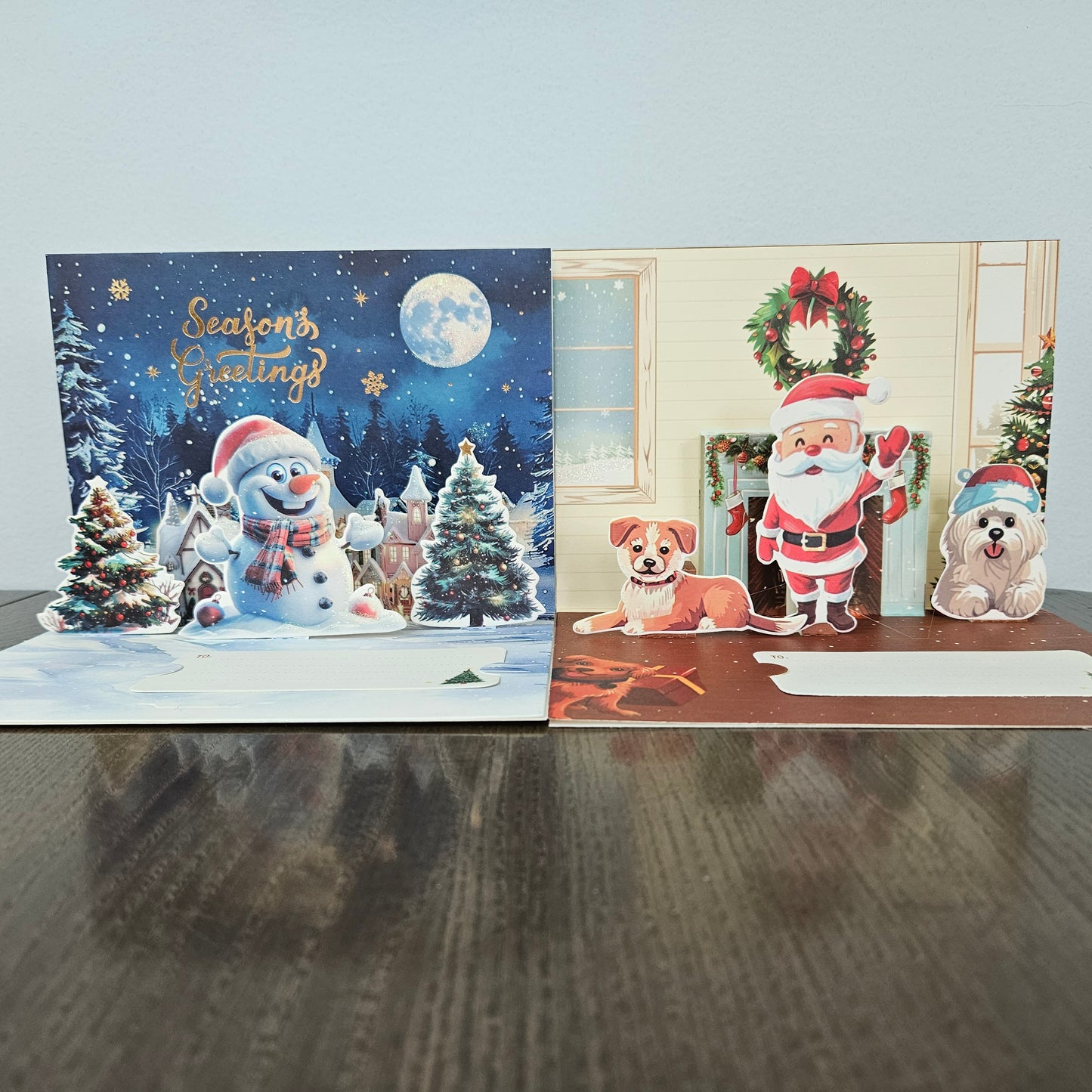 Two Christmas pop-up greeting cards displayed side by side on a dark wooden table against a light gray wall. The left card features a snowy night scene with a snowman, Christmas trees, and the text 'Season's Greetings'.