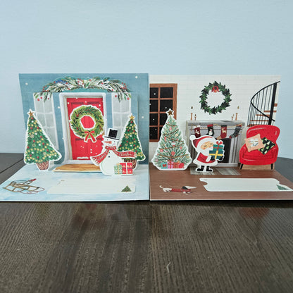 A Christmas-themed pop-up cardboard card set displayed on a dark wooden table against a light gray wall. The set includes a pop-up scene of Santa Claus at a fireplace with a red armchair and Christmas tree, and another pop-up scene of a festive front door with a wreath, snowman, and Christmas trees.