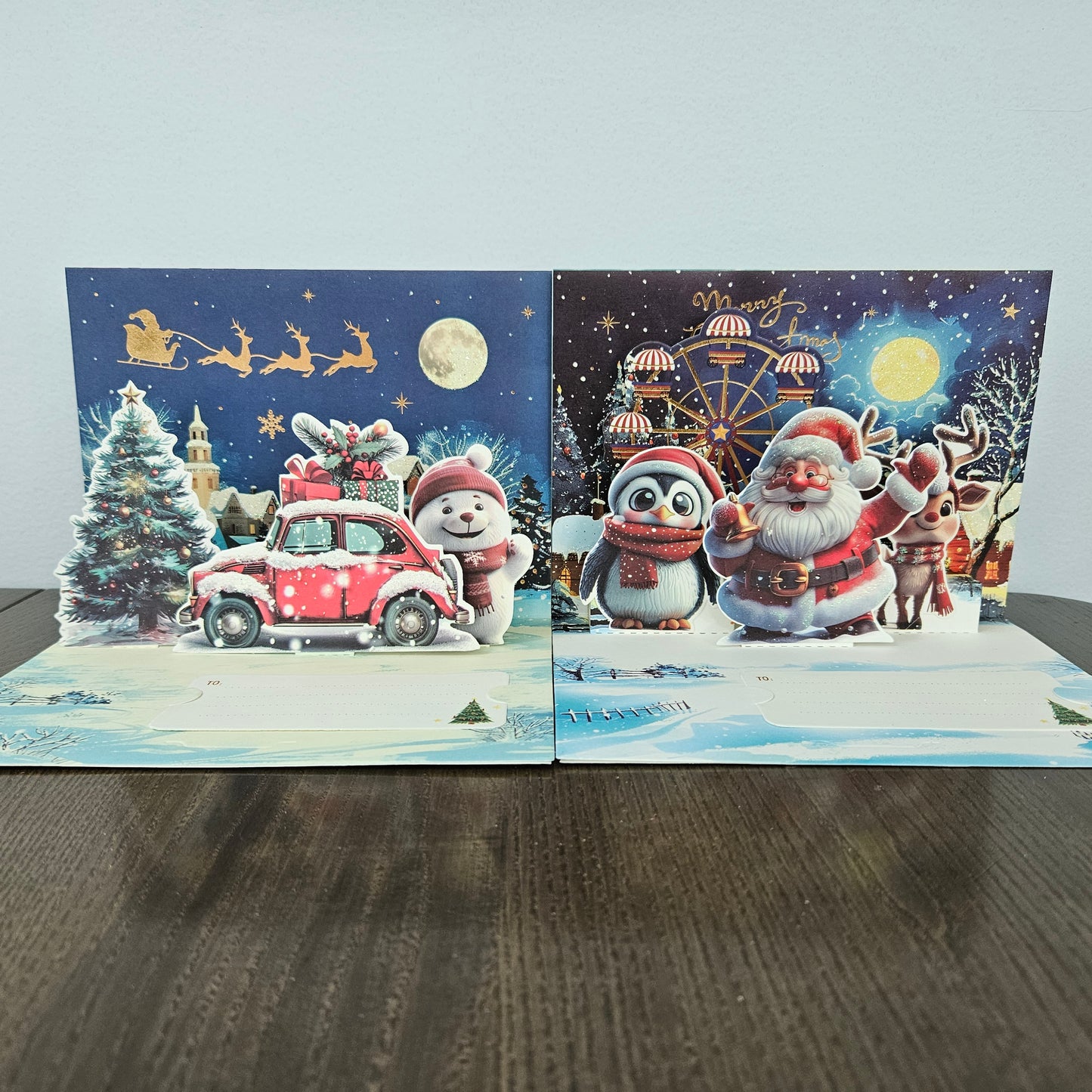 Two 3D pop-up Christmas greeting cards displayed side-by-side on a dark wooden table against a light gray wall. The left card features a snowy night scene with Santa Claus in a red car, a snowman, a Christmas tree, and sleighs.