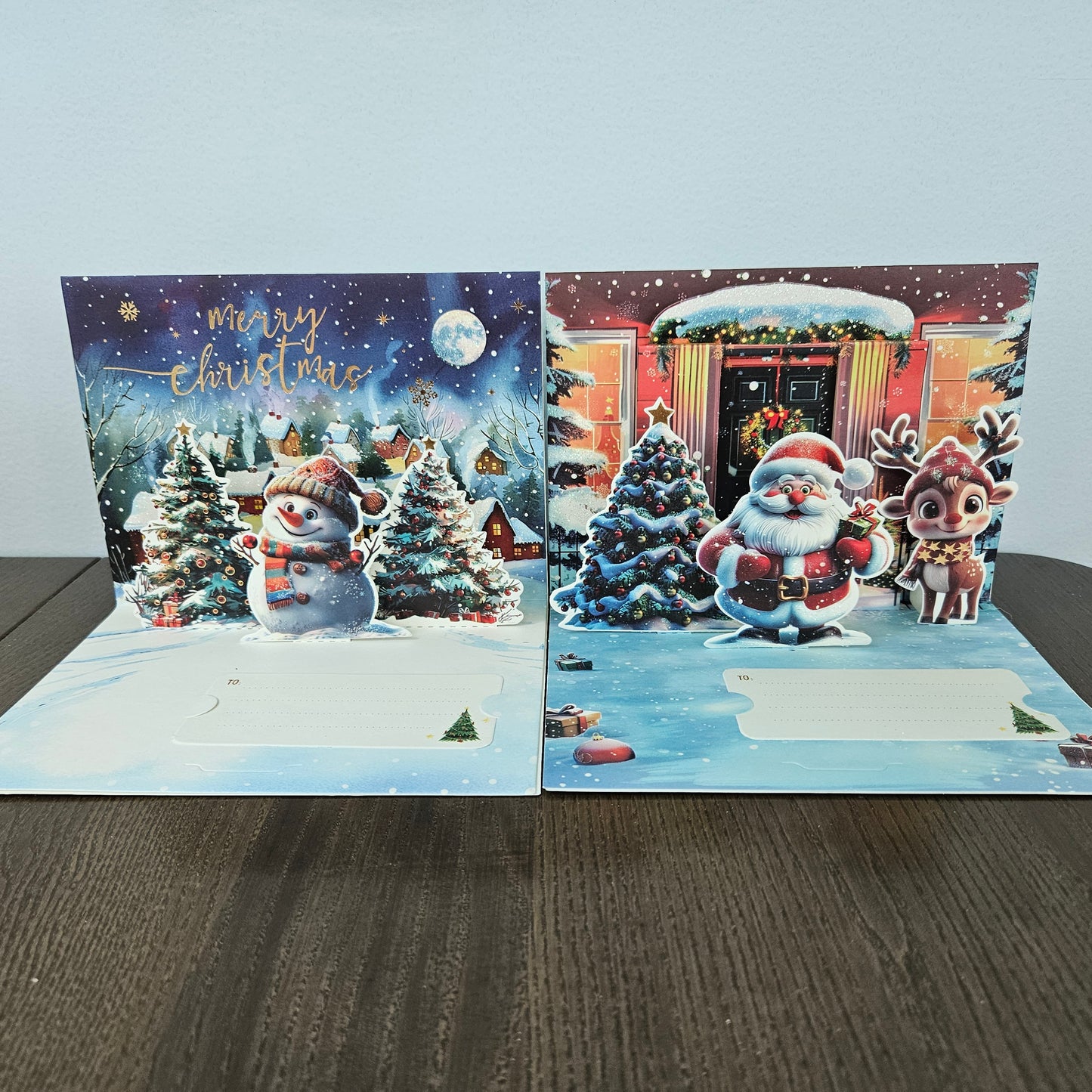Two 3D pop-up Christmas greeting cards displayed side-by-side on a dark wooden table against a light gray wall. The left card features a snowy night scene with a snowman, decorated trees, and 'Merry Christmas' in gold script.