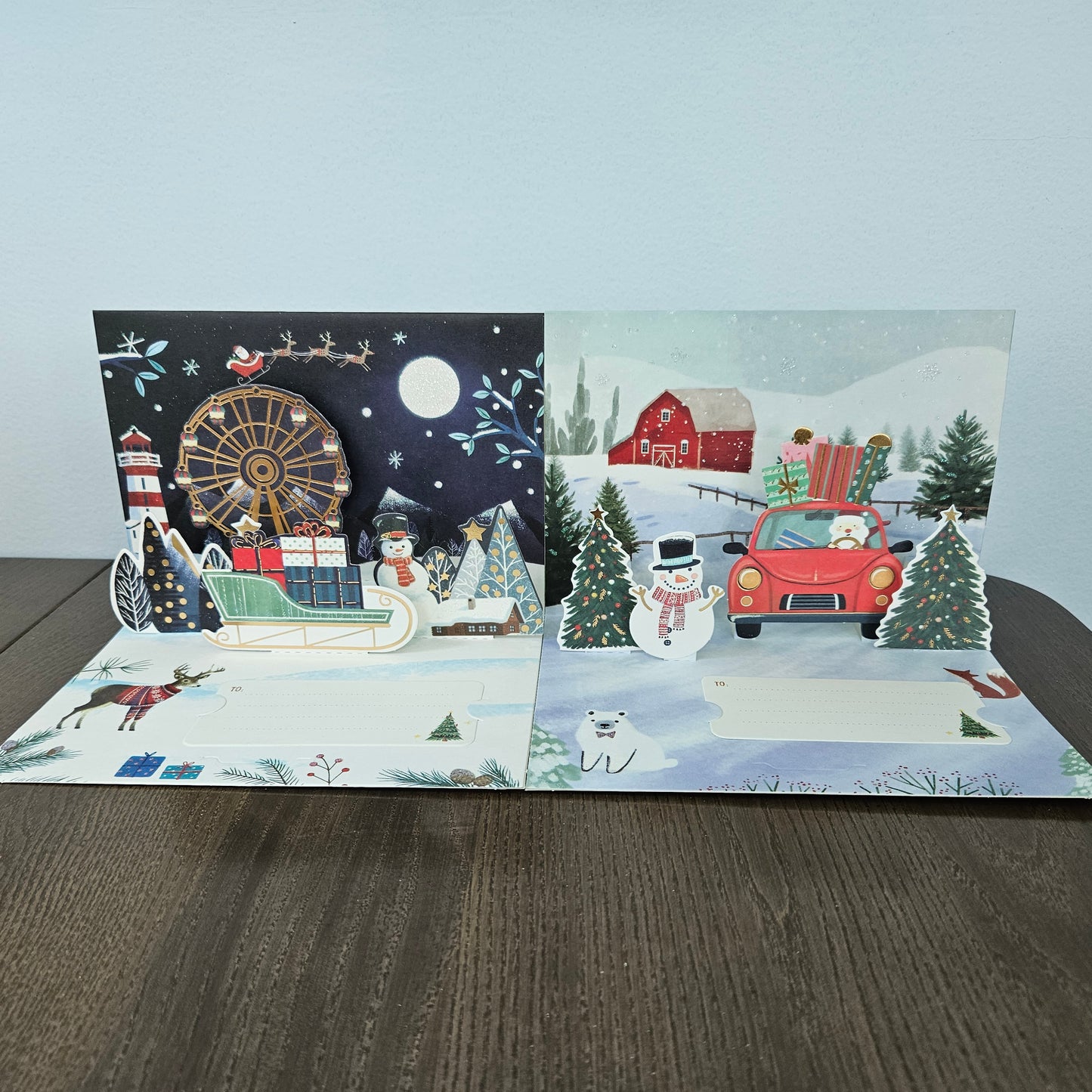 A 3D pop-up cardboard Christmas greeting card displayed open on a dark wooden table against a light gray wall. The card features two distinct pop-up scenes: the left side shows a festive night scene with Santa in a sleigh, a lighthouse, a Ferris wheel, and a lighthouse pop-up; the right side shows a snowy winter landscape with a red barn, a red car carrying a Christmas tree, a snowman, and a polar bear pop-up.