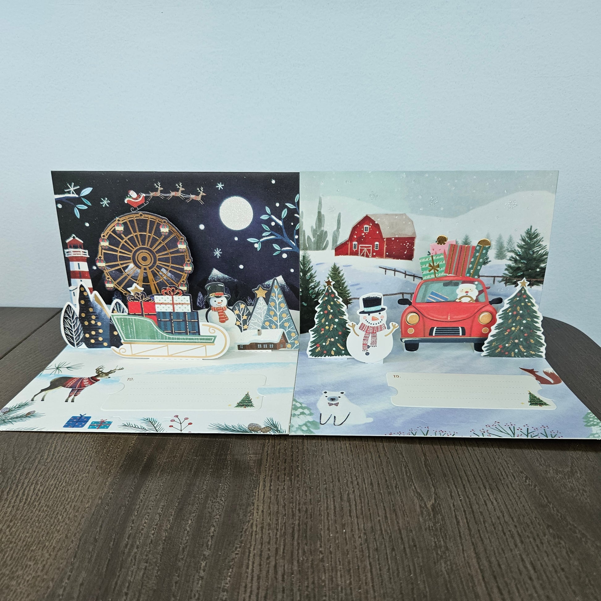 A 3D pop-up cardboard Christmas greeting card displayed open on a dark wooden table against a light gray wall. The card features two distinct pop-up scenes: the left side shows a festive night scene with Santa in a sleigh, a lighthouse, a Ferris wheel, and a lighthouse pop-up; the right side shows a snowy winter landscape with a red barn, a red car carrying a Christmas tree, a snowman, and a polar bear pop-up.