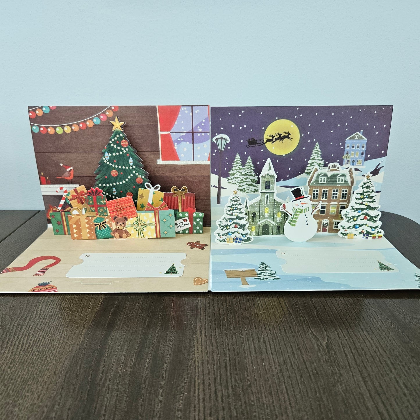 Two open 3D pop-up Christmas greeting cards displayed side-by-side on a dark wooden table against a light gray wall. The left card features a festive Christmas tree, wrapped gifts, and a cozy window scene.