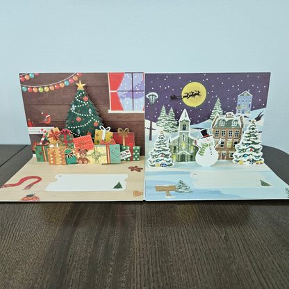 Two open 3D pop-up Christmas greeting cards displayed side-by-side on a dark wooden table against a light gray wall. The left card features a festive Christmas tree, wrapped gifts, and a cozy window scene.