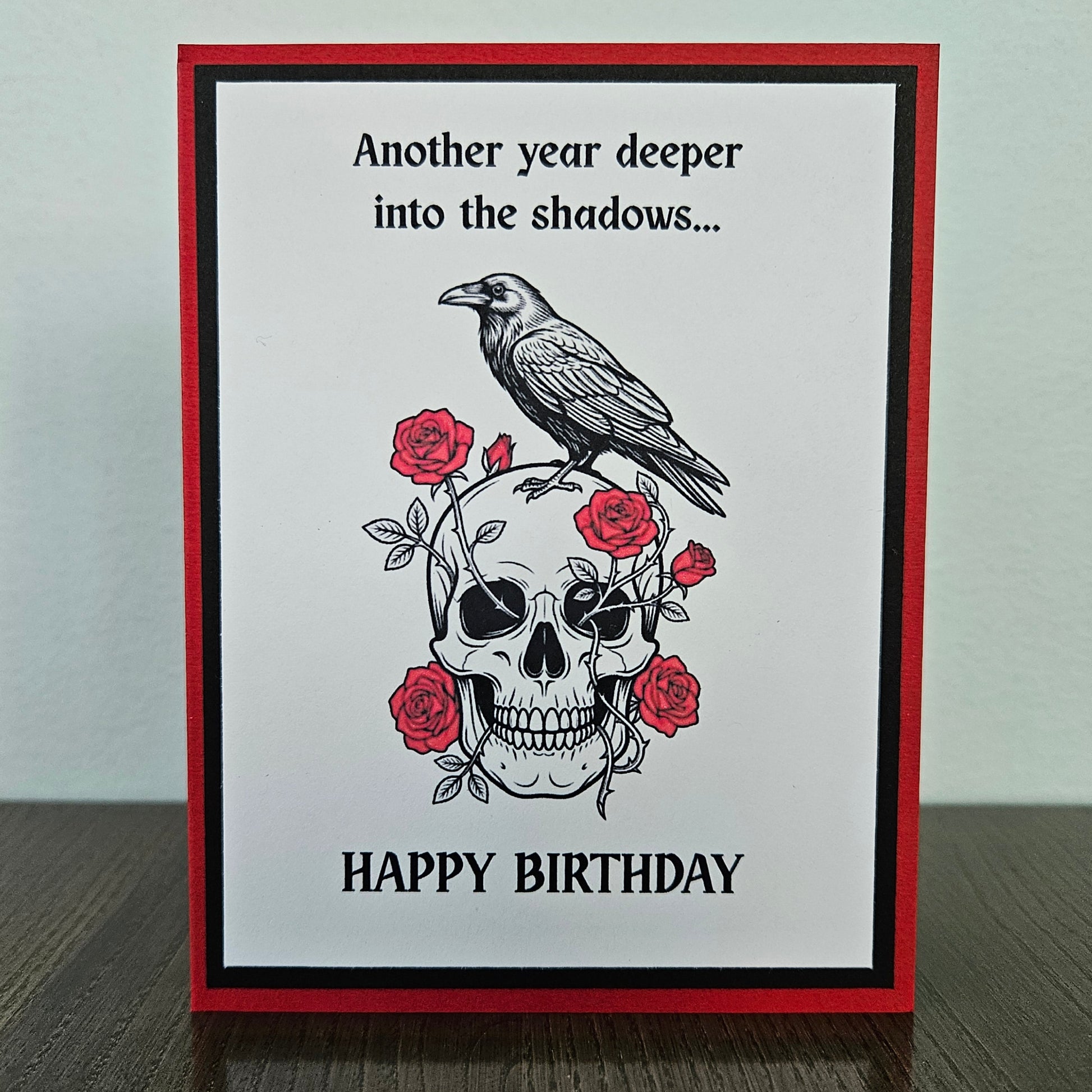 Front of a handmade A2 gothic birthday card featuring layered red, black, and white cardstock with a raven perched on a skull wrapped in red roses with text reading Another year deeper into the shadows... HAPPY BIRTHDAY.