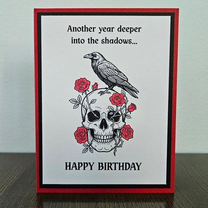 Front of a handmade A2 gothic birthday card featuring layered red, black, and white cardstock with a raven perched on a skull wrapped in red roses with text reading Another year deeper into the shadows... HAPPY BIRTHDAY.