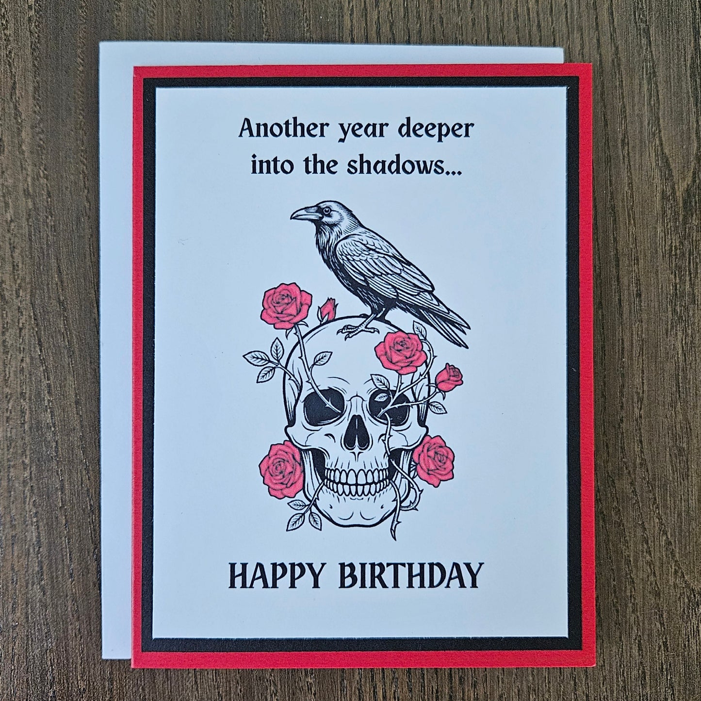 Front of a handmade A2 gothic birthday card featuring layered red, black, and white cardstock with a raven perched on a skull wrapped in red roses with text reading Another year deeper into the shadows... HAPPY BIRTHDAY.