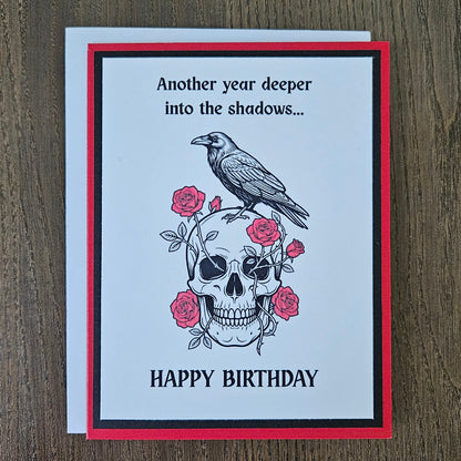 Front of a handmade A2 gothic birthday card featuring layered red, black, and white cardstock with a raven perched on a skull wrapped in red roses with text reading Another year deeper into the shadows... HAPPY BIRTHDAY.