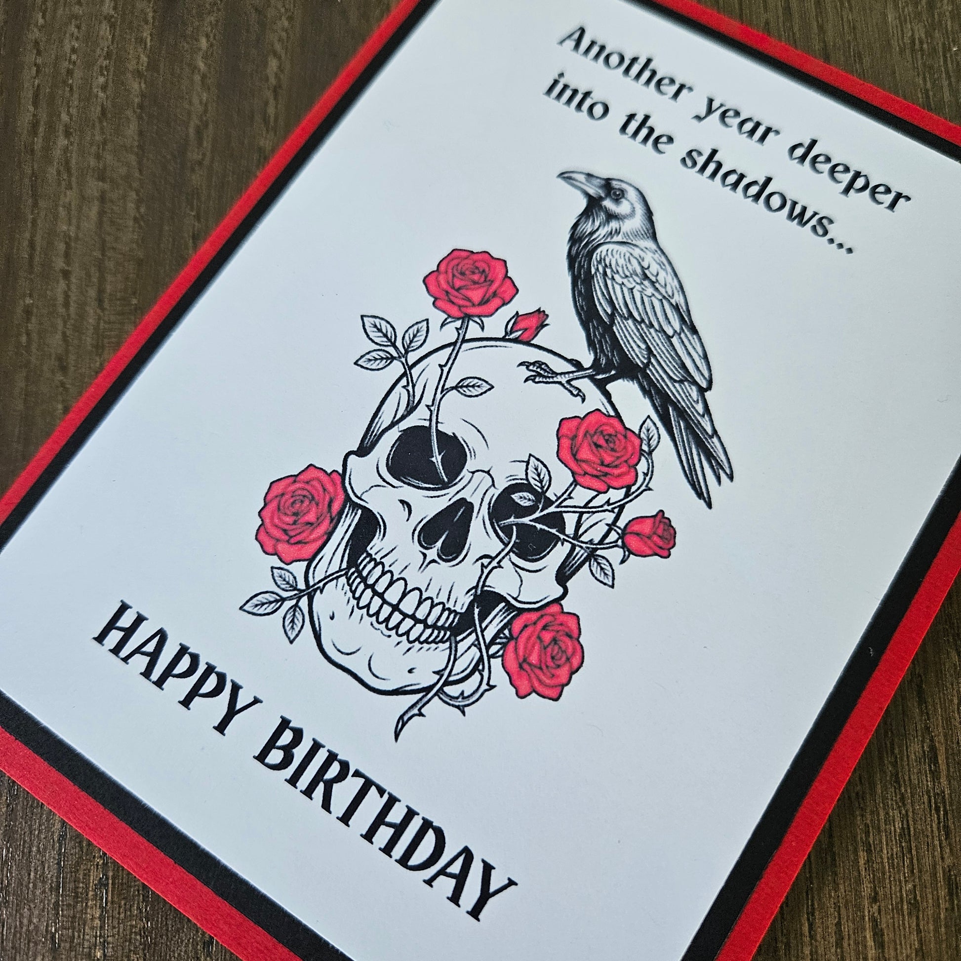 A square greeting card with a white center, red border, and black text reading 'Another year deeper into the shadows.