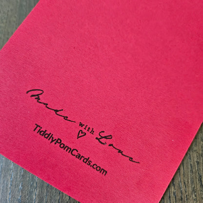 A close-up of a pinkish-red textured card or paper with black printed text reading 'Made with Love' and 'TiddlyPornCards.com' on a wood surface.