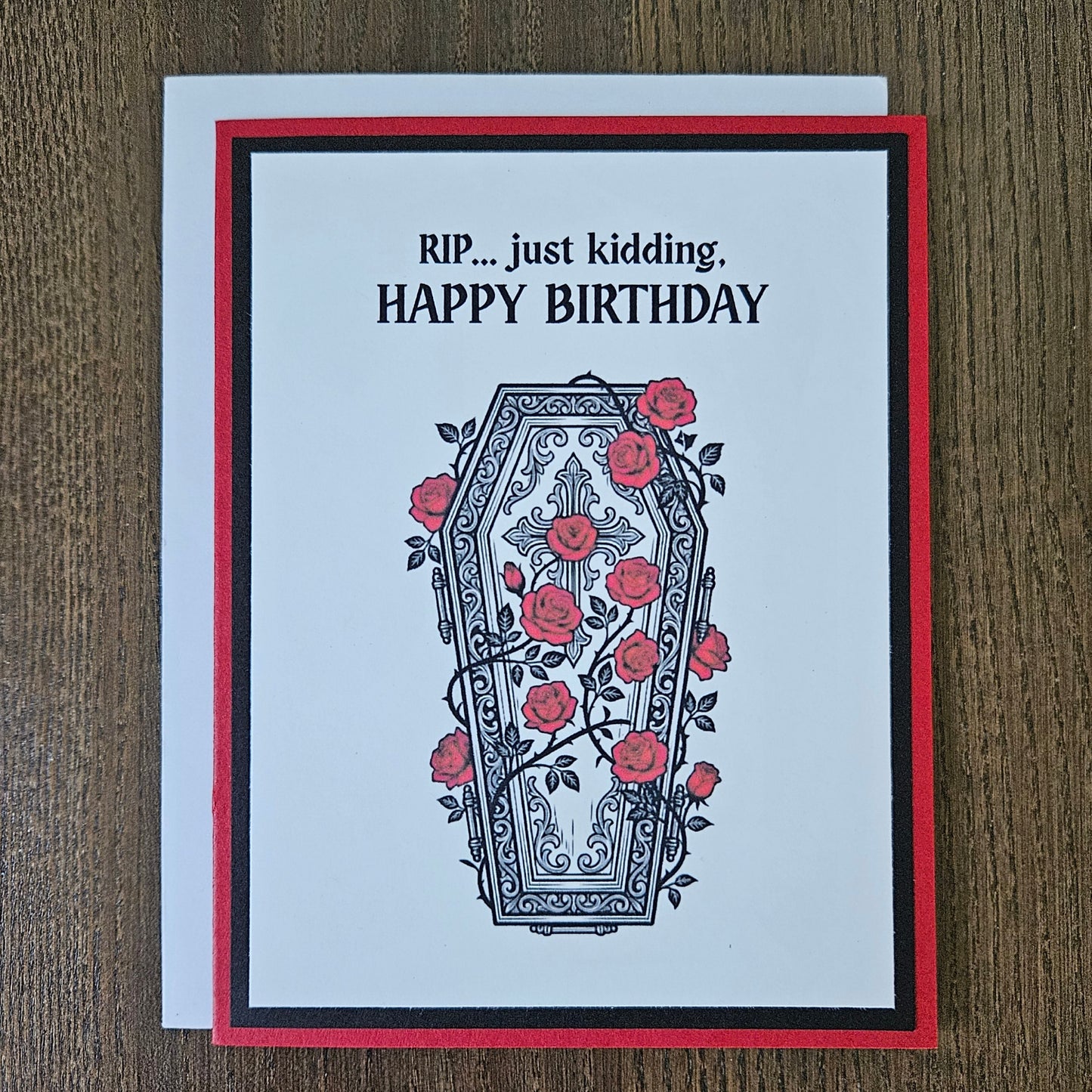 Front of a handmade A2 gothic birthday card featuring a layered design of red, black, and white cardstock. Above an ornate illustration of a coffin covered in red roses and vines is the snarky sentiment: RIP... just kidding, HAPPY BIRTHDAY.