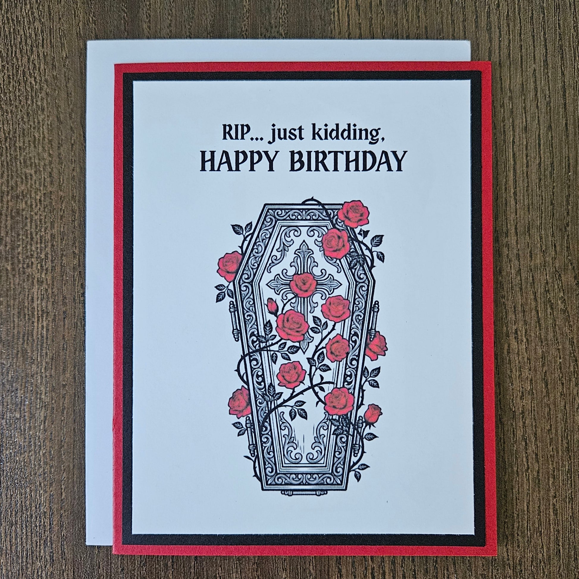 Front of a handmade A2 gothic birthday card featuring a layered design of red, black, and white cardstock. Above an ornate illustration of a coffin covered in red roses and vines is the snarky sentiment: RIP... just kidding, HAPPY BIRTHDAY.