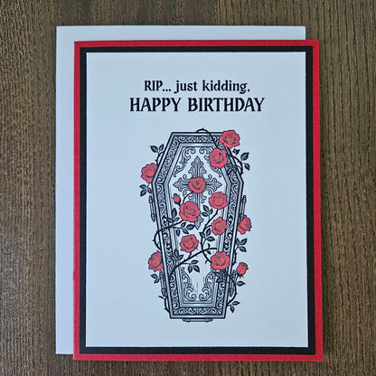 Front of a handmade A2 gothic birthday card featuring a layered design of red, black, and white cardstock. Above an ornate illustration of a coffin covered in red roses and vines is the snarky sentiment: RIP... just kidding, HAPPY BIRTHDAY.