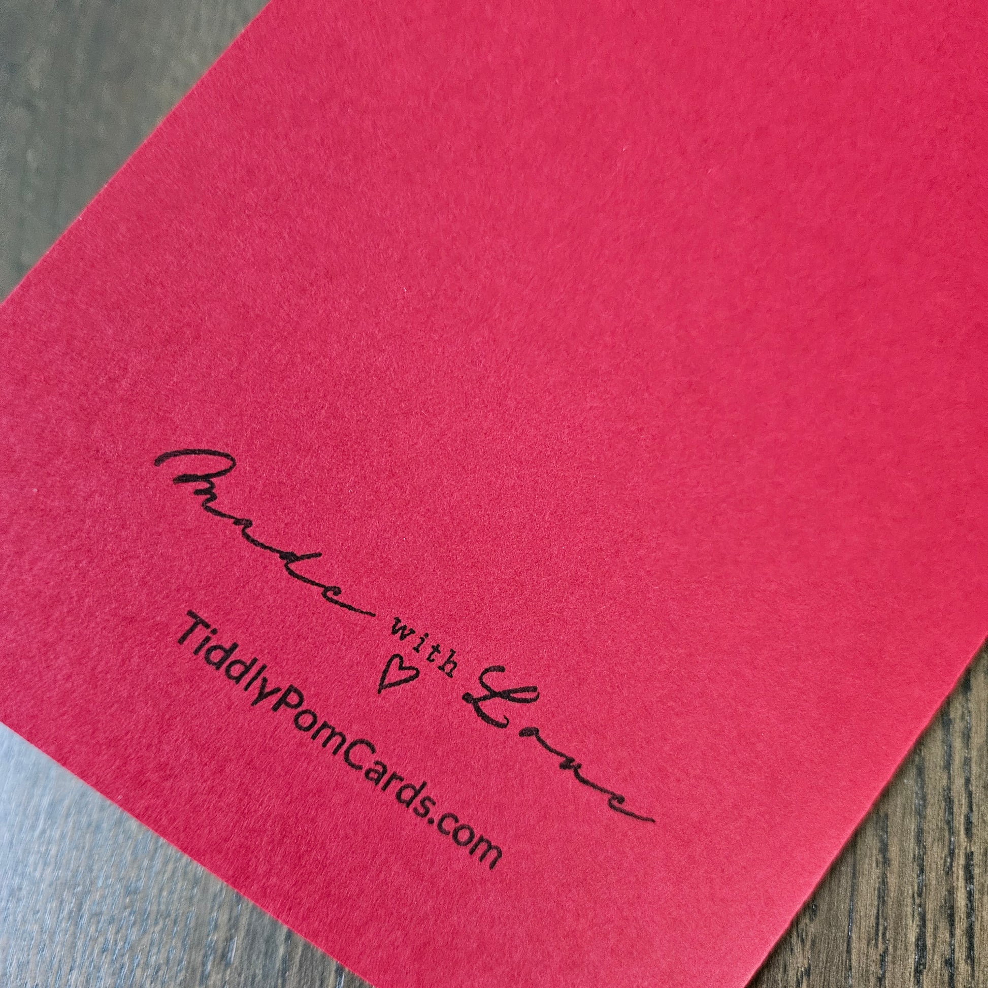 A close-up of a red textured card or paper with black printed text reading 'Made with Love' and 'TiddlyPomCards.com' on a wood surface.