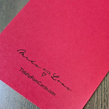 A close-up of a red textured card or paper with black printed text reading 'Made with Love' and 'TiddlyPomCards.com' on a wood surface.