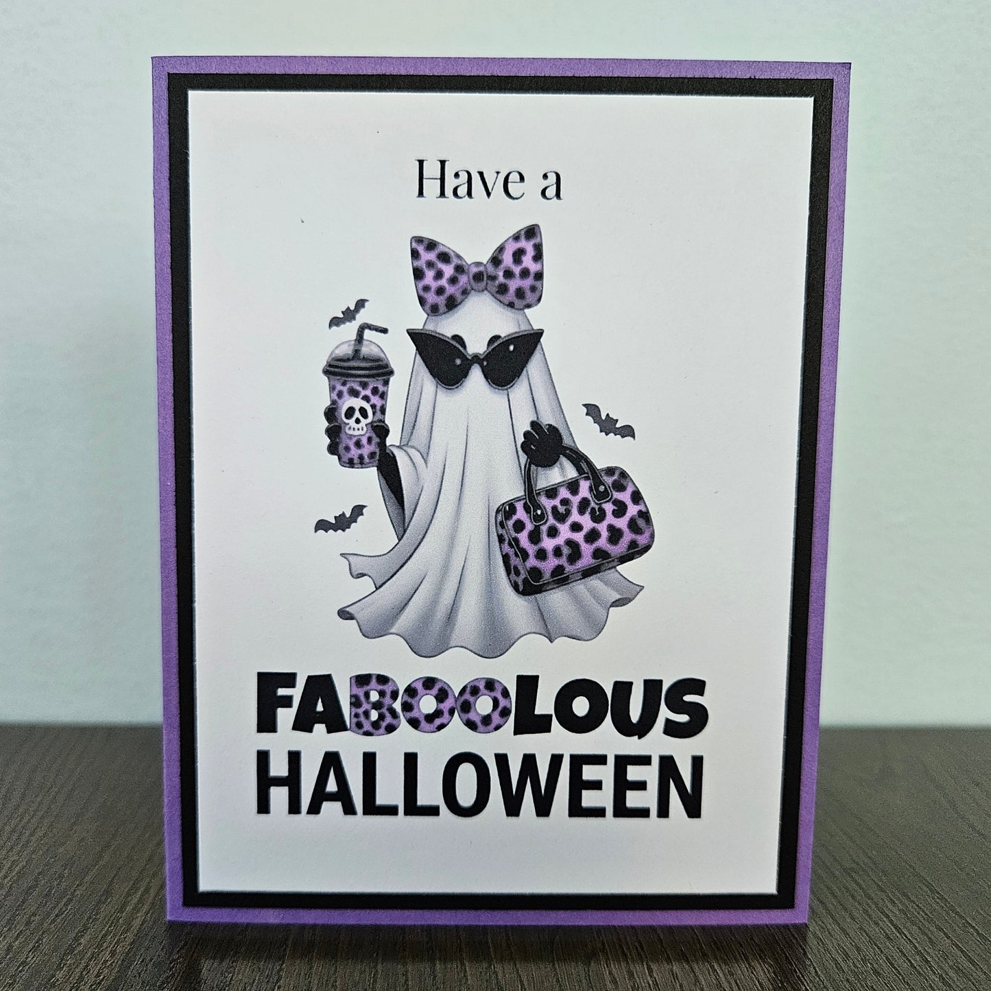 Front of a handmade A2 Halloween card featuring a layered purple and black border around a fashionable ghost wearing a purple leopard bow and sunglasses, holding an iced coffee and purse, with text reading Have a FABOOLOUS HALLOWEEN.
