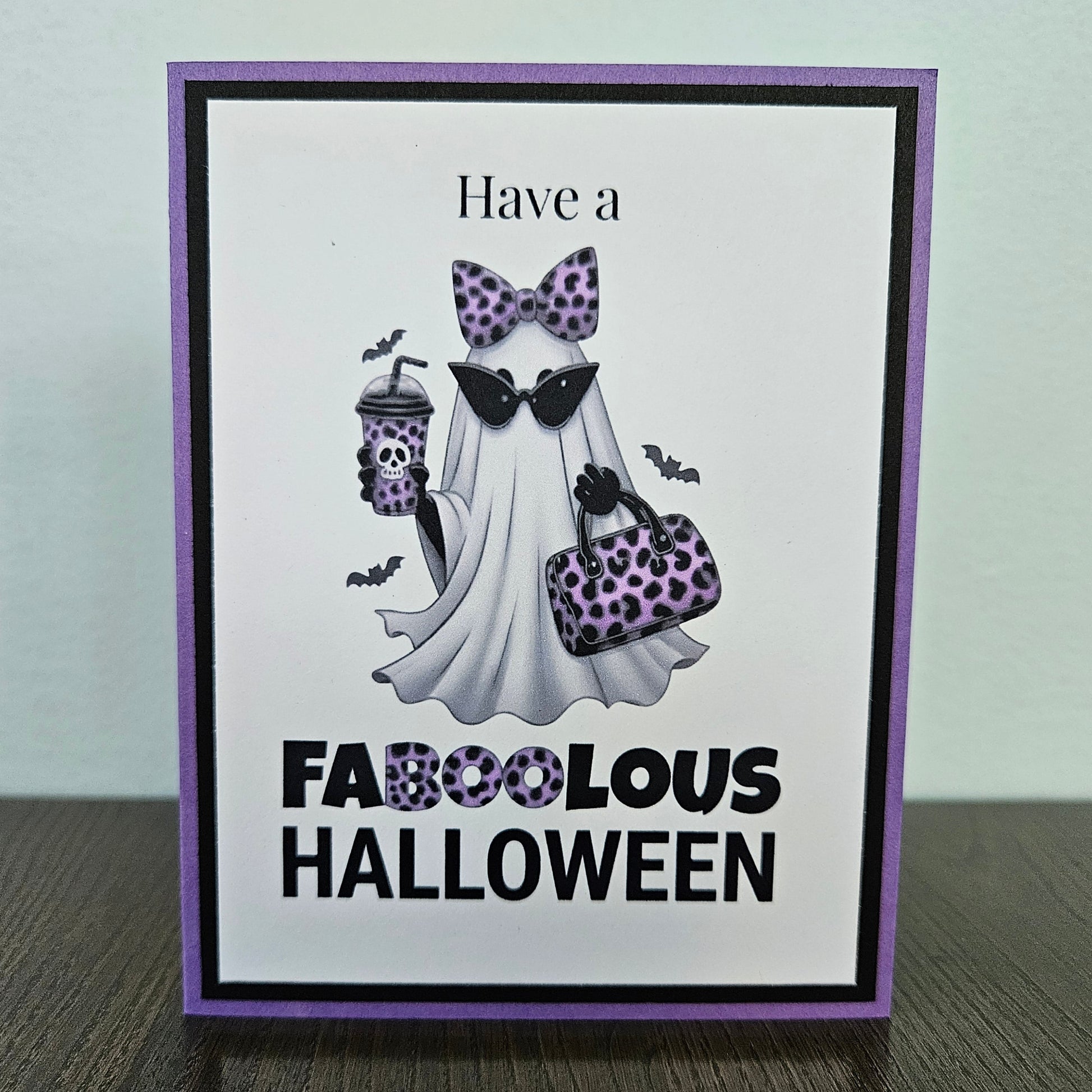Front of a handmade A2 Halloween card featuring a layered purple and black border around a fashionable ghost wearing a purple leopard bow and sunglasses, holding an iced coffee and purse, with text reading Have a FABOOLOUS HALLOWEEN.