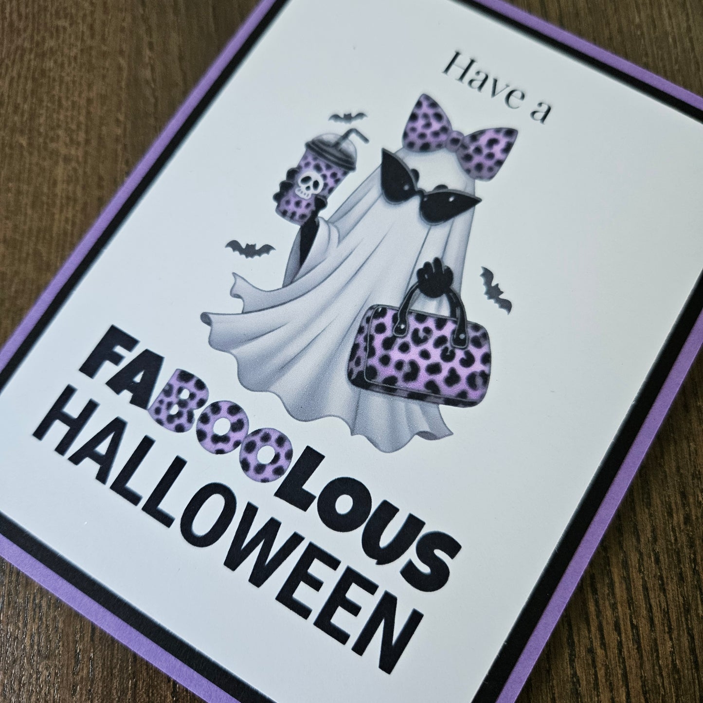A square printed card or sign with a Halloween-themed illustration and the text 'Have a FABULOUS HALLOWEEN,' placed on a wooden surface. The design shows a white ghost with purple leopard-print accents, black bats, and bold black lettering.