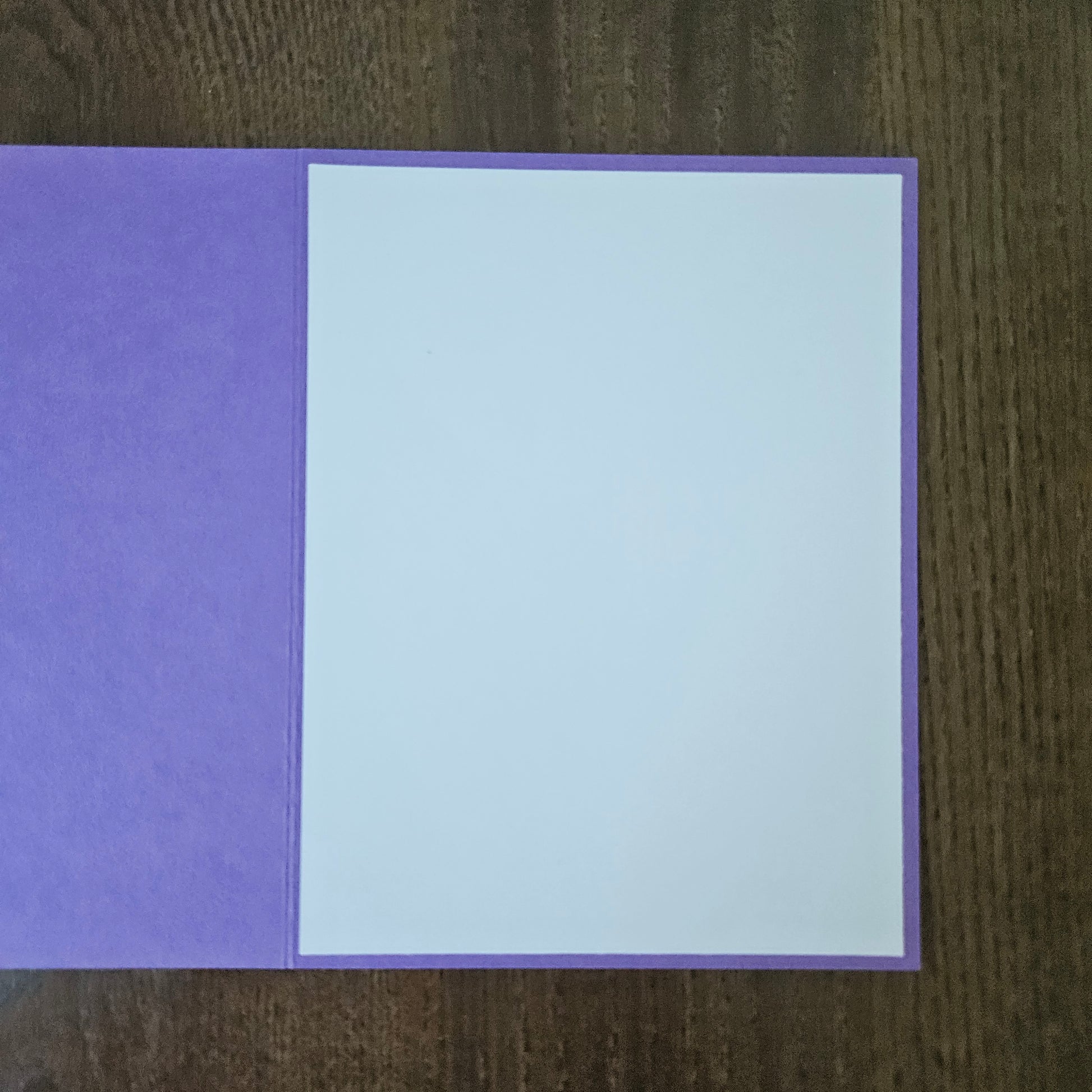 A purple greeting card with a blank white insert, displayed flat on a dark brown wooden surface.