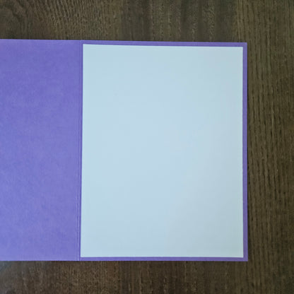 A purple greeting card with a blank white insert, displayed flat on a dark brown wooden surface.