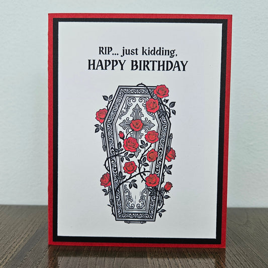 Front of a handmade A2 gothic birthday card featuring a layered design of red, black, and white cardstock. Above an ornate illustration of a coffin covered in red roses and vines is the snarky sentiment: RIP... just kidding, HAPPY BIRTHDAY.