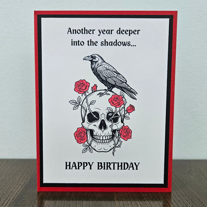 Front of a handmade A2 gothic birthday card featuring layered red, black, and white cardstock with a raven perched on a skull wrapped in red roses with text reading Another year deeper into the shadows... HAPPY BIRTHDAY.