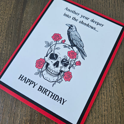 Front of a handmade A2 gothic birthday card featuring layered red, black, and white cardstock with a raven perched on a skull wrapped in red roses with text reading Another year deeper into the shadows... HAPPY BIRTHDAY.