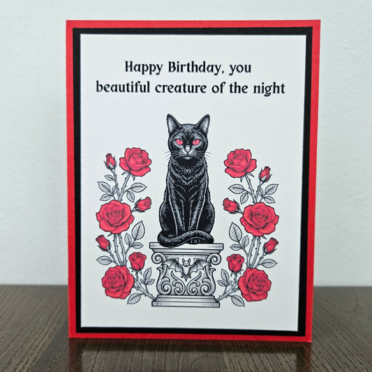 Front of a handmade A2 gothic birthday card featuring a black cat with red eyes sitting on a pedestal surrounded by red roses with the text Happy Birthday, you beautiful creature of the night.