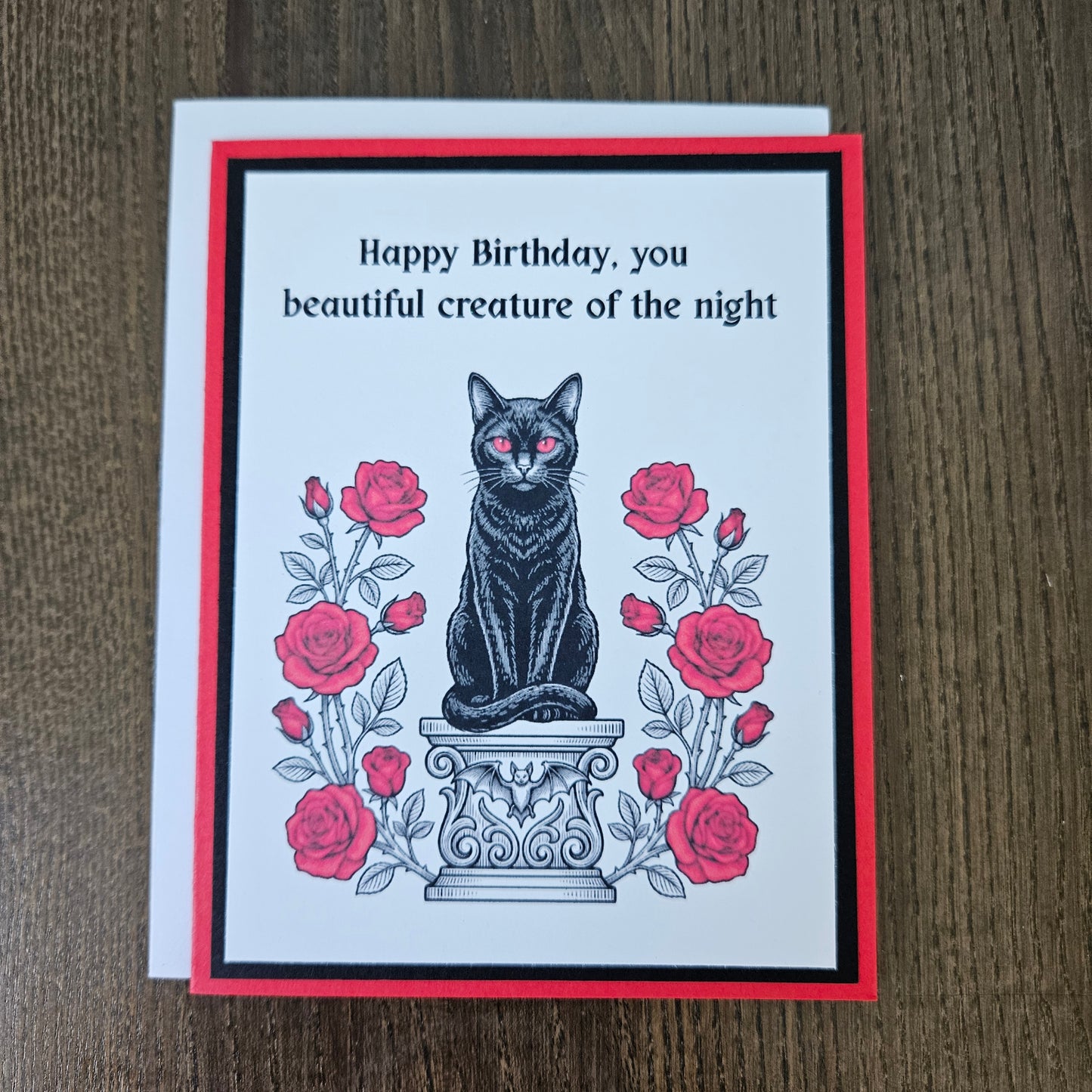 Front of a handmade A2 gothic birthday card featuring a black cat with red eyes sitting on a pedestal surrounded by red roses with the text Happy Birthday, you beautiful creature of the night.