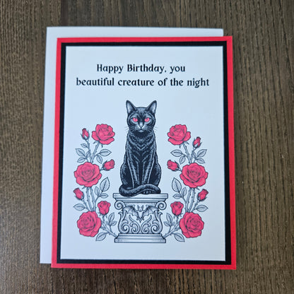 Front of a handmade A2 gothic birthday card featuring a black cat with red eyes sitting on a pedestal surrounded by red roses with the text Happy Birthday, you beautiful creature of the night.