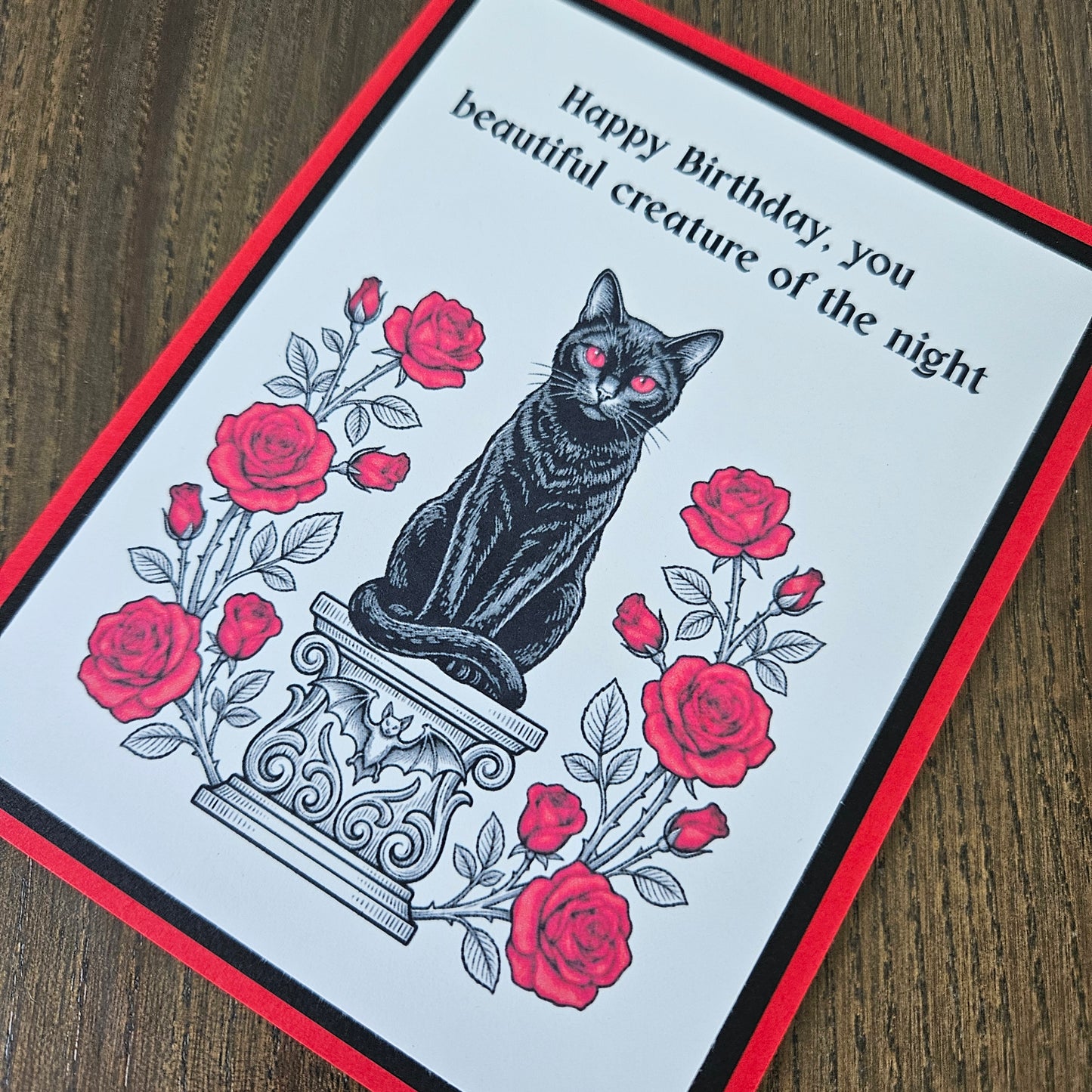 Front of a handmade A2 gothic birthday card featuring a black cat with red eyes sitting on a pedestal surrounded by red roses with the text Happy Birthday, you beautiful creature of the night.