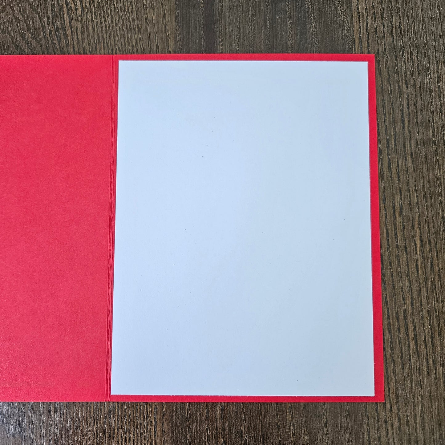 A red greeting card or paper envelope with a large blank white center panel, photographed flat on a dark wood surface.