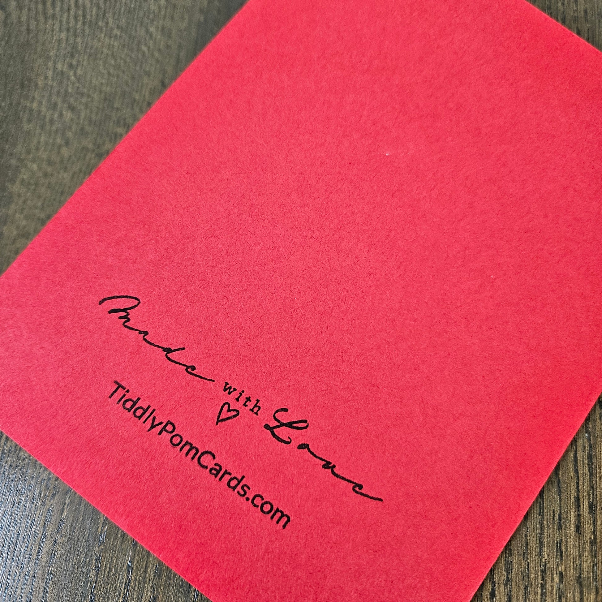 A red square pom card with black printed text reading 'Made with Love' and 'TidilyPomCards.com', placed on a dark wood surface.