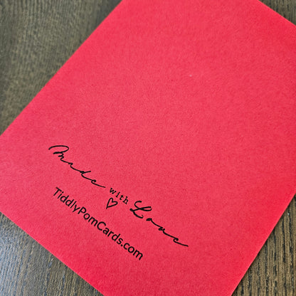 A red square pom card with black printed text reading 'Made with Love' and 'TidilyPomCards.com', placed on a dark wood surface.