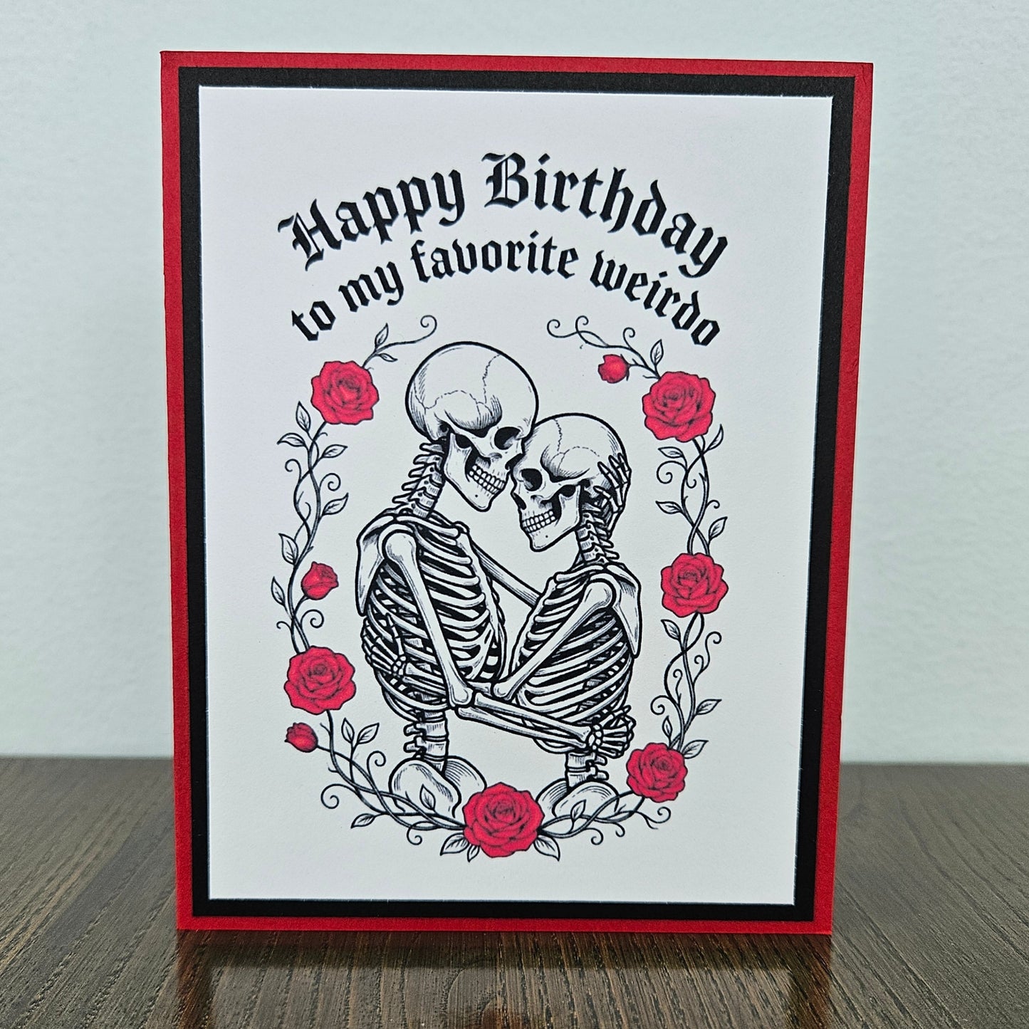 Front of a handmade A2 gothic birthday card featuring layered red and black borders around an illustration of two skeletons embracing within a wreath of red roses with text reading Happy Birthday to my favorite weirdo.