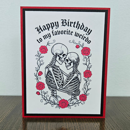 Front of a handmade A2 gothic birthday card featuring layered red and black borders around an illustration of two skeletons embracing within a wreath of red roses with text reading Happy Birthday to my favorite weirdo.