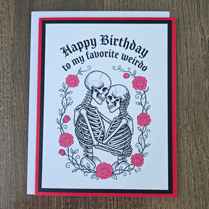 Front of a handmade A2 gothic birthday card featuring layered red and black borders around an illustration of two skeletons embracing within a wreath of red roses with text reading Happy Birthday to my favorite weirdo.