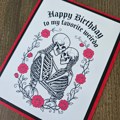 A handmade greeting card featuring a black and white illustration of a skeleton embracing a skull, surrounded by red roses and the text 'Happy Birthday to my favorite weirdo'. The card has a white center, a black and red border, and is photographed on a wooden surface.