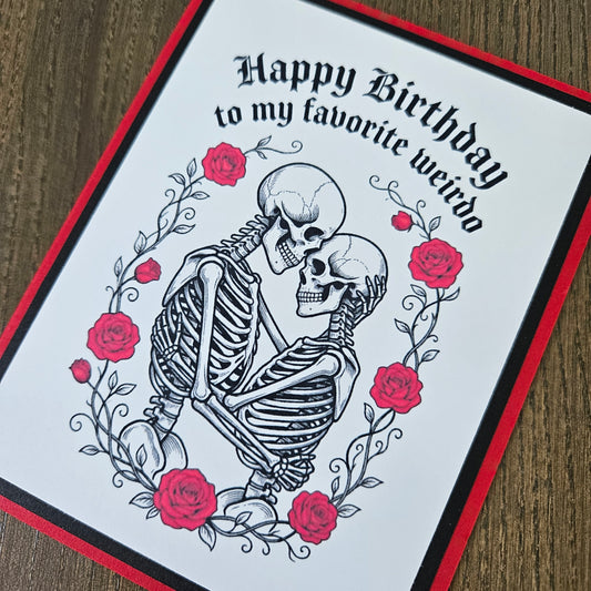 A handmade greeting card featuring a black and white illustration of a skeleton embracing a skull, surrounded by red roses and the text 'Happy Birthday to my favorite weirdo'. The card has a white center, a black and red border, and is photographed on a wooden surface.