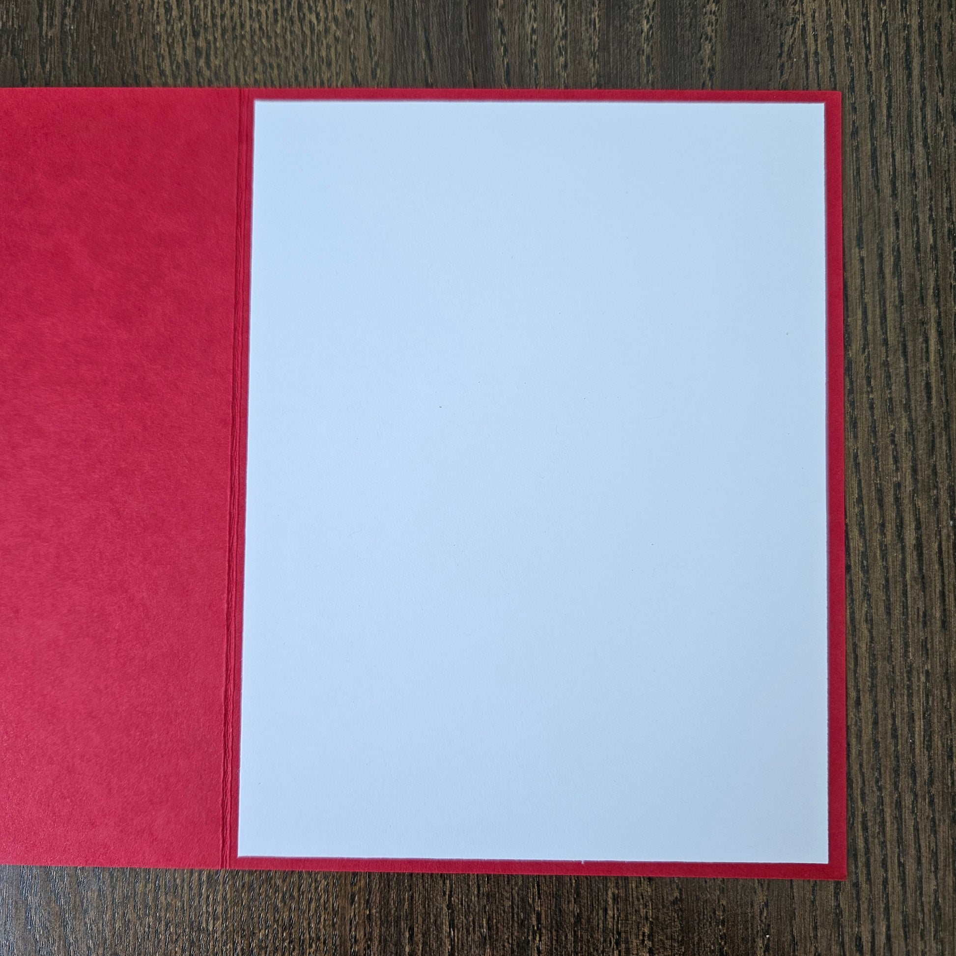 A red greeting card or envelope with a large blank white center panel, photographed flat on a dark wood surface.