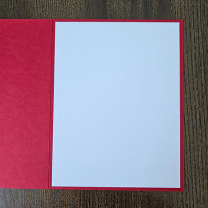 A red greeting card or envelope with a large blank white center panel, photographed flat on a dark wood surface.