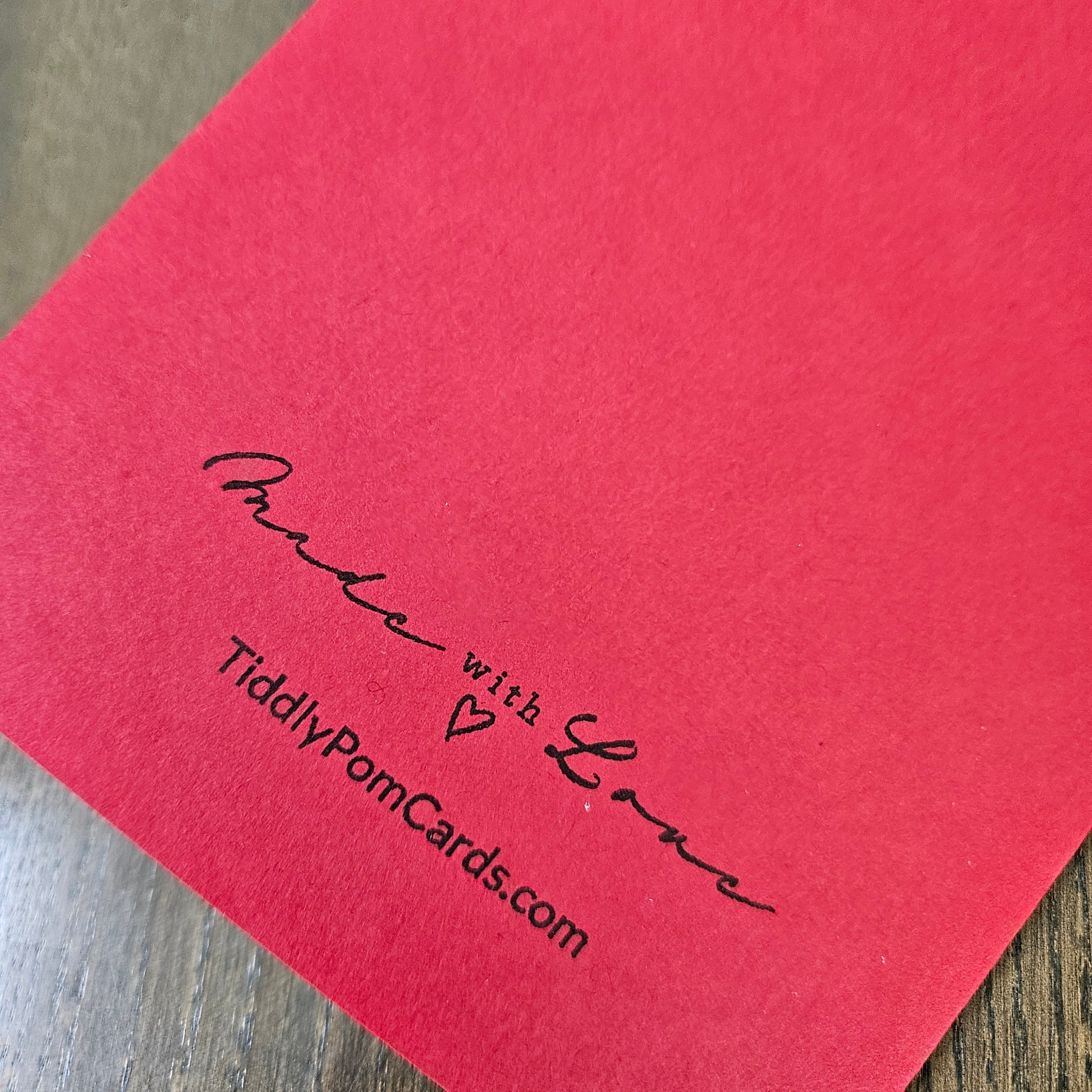 A close-up of a red card with black printed text reading 'Made with Love' and 'TiddlyPomCards.com' on a wooden surface.