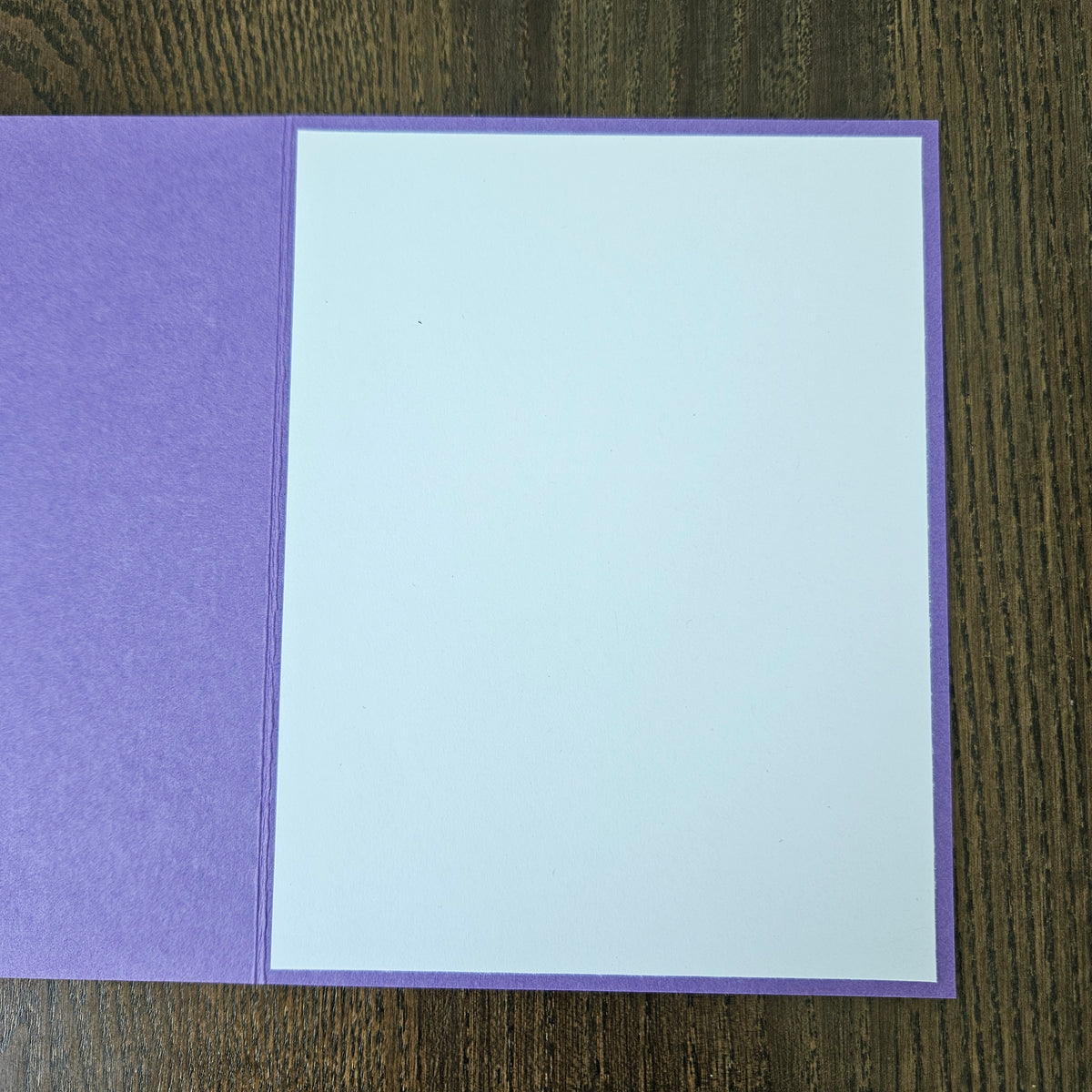 A square purple card or envelope with a large blank white insert, photographed from above on a dark wood surface.
