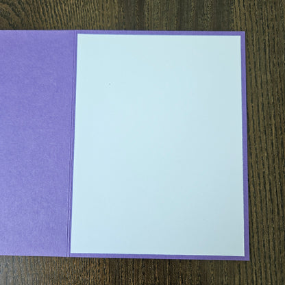 A square purple card or envelope with a large blank white insert, photographed from above on a dark wood surface.