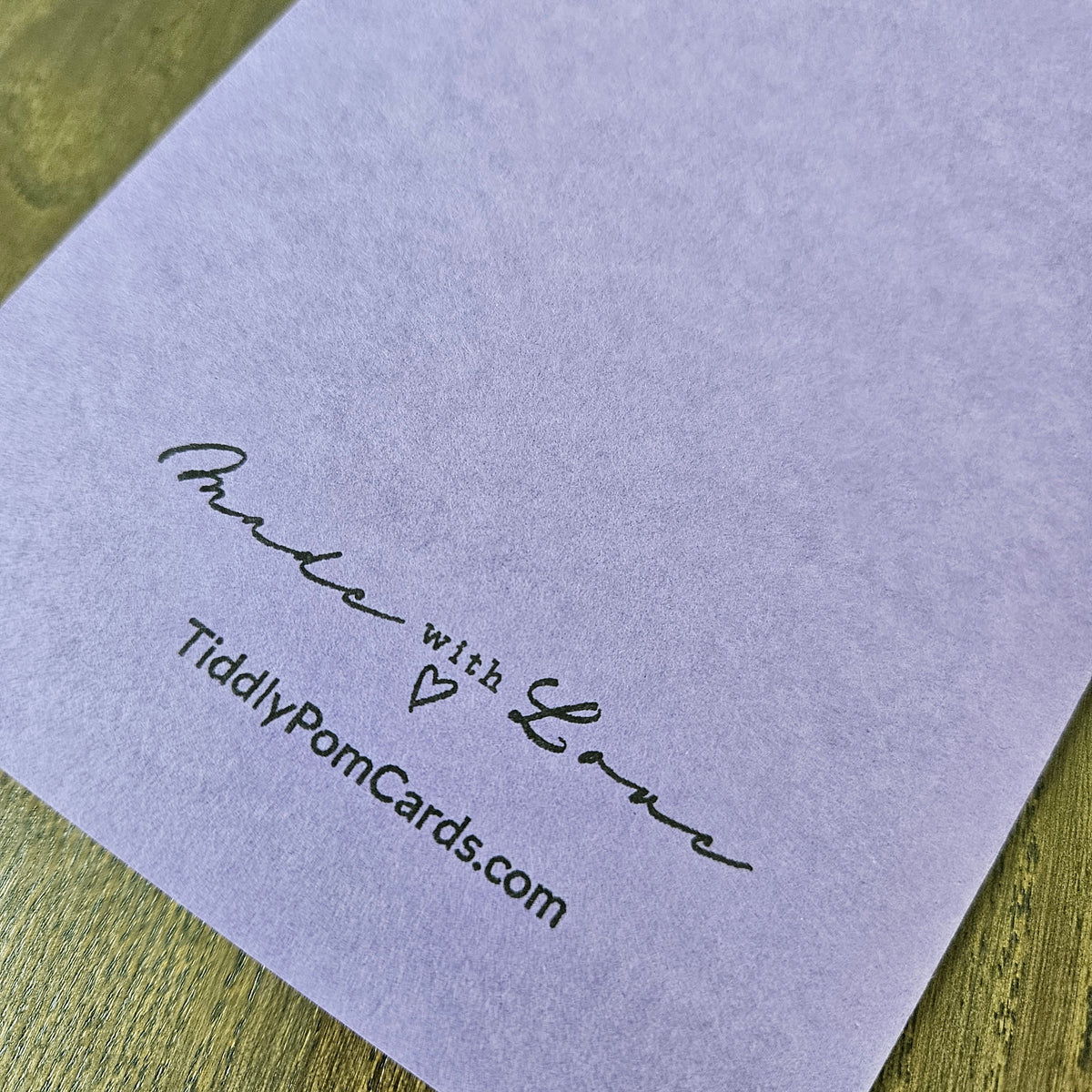 A close-up of a light lavender paper card printed with the words 'Made with Love' in black script and 'TiddlyPomCards.com' in black text, placed on a wooden surface.