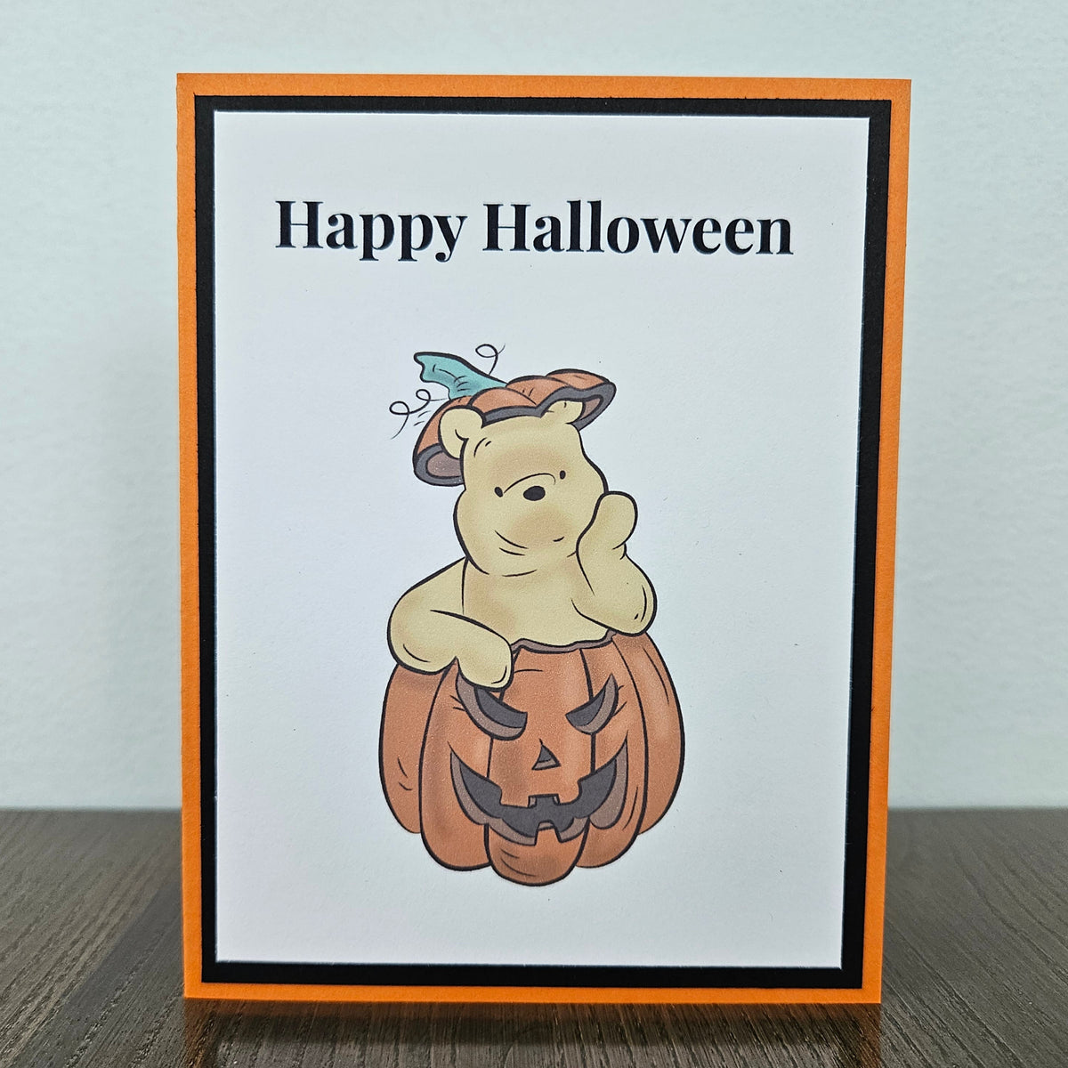 A handmade Halloween greeting card featuring Winnie the Pooh holding a carved pumpkin, with the text 'Happy Halloween' at the top. The card has an orange border, a black inner border, and is photographed standing on a dark wooden surface against a light gray wall.