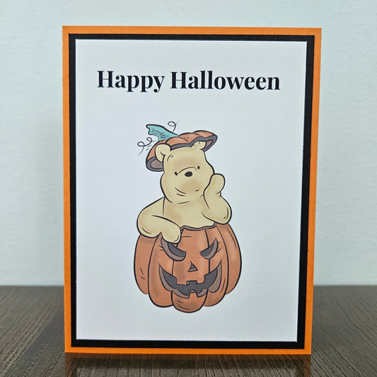 A handmade Halloween greeting card featuring Winnie the Pooh holding a carved pumpkin, with the text 'Happy Halloween' at the top. The card has an orange border, a black inner border, and is photographed standing on a dark wooden surface against a light gray wall.